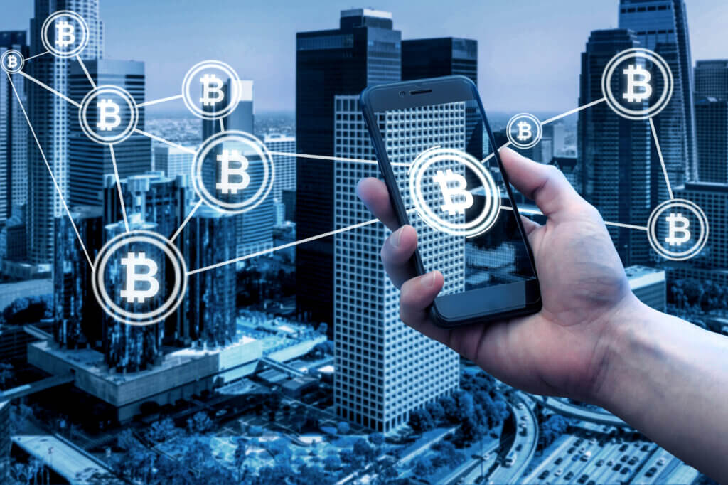Bitcoin... ¿en el sector inmobiliario? Celular muestra el símbolo de bitcoin y de fondo edificios y propiedades inmobiliarias.